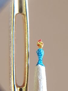 Gelber Fisch auf blauem Gund in Zahnstocher geschnitten neben einem Nadelöhr als Größenvergleich. Das Nadelör ist um ein vielfaches Größer als die Fischskulptur